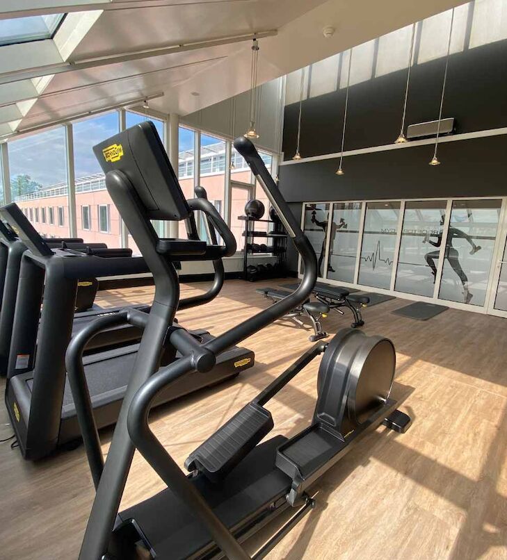 Fitnessstudio mit Ellipsentrainer im Vordergrund, Laufbändern im Hintergrund und Glasfenstern.