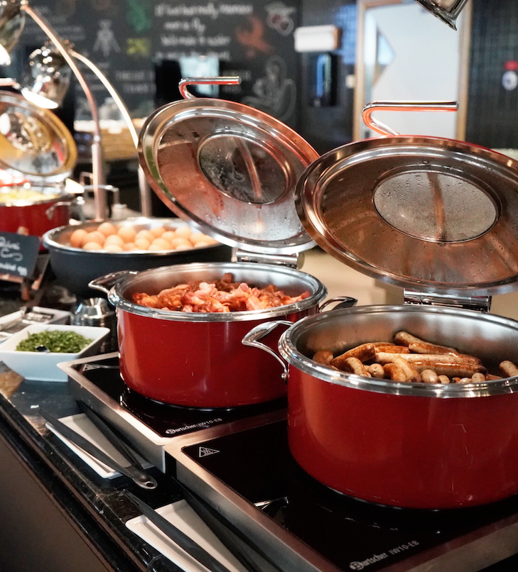 Ein Hotel-Frühstücksbuffet mit roten Töpfen auf Wärmeplatten, in denen Würstchen und Kartoffeln stehen.