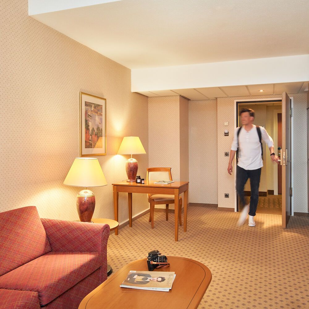 Ein Mann betritt ein Hotelzimmer mit Rucksack. Das Zimmer ist mit einem Schreibtisch, Lampen und einem Sofa ausgestattet.