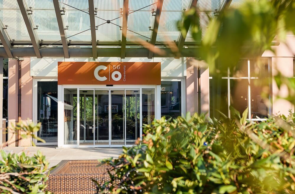 Eingang zu einem Gebäude mit der Aufschrift „Co“, orangefarbenen Schildern, Glastüren und einem überdachten Gehweg.