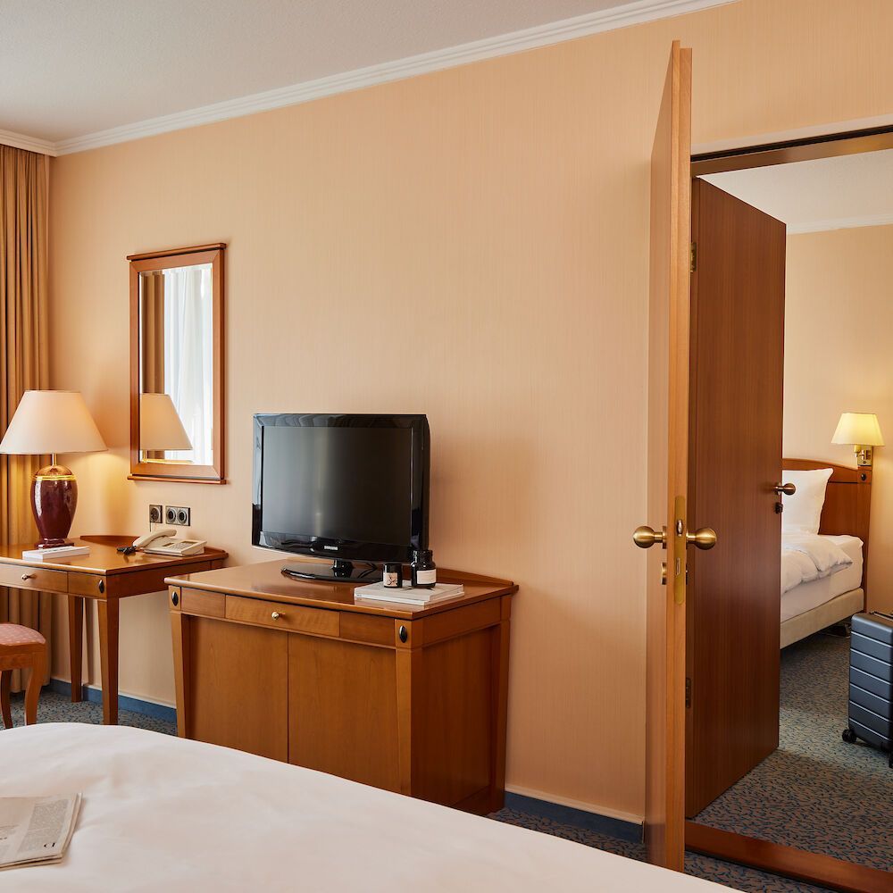 Hotelzimmer mit Bett, Fernseher, Schreibtisch und einer teilweise geöffneten Tür zu einem weiteren Zimmer mit Bett.