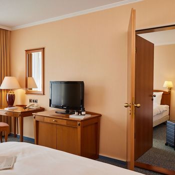 Hotelzimmer-Interieur mit Bett, Fernseher, Schreibtisch und einer teilweise geöffneten Tür zu einem weiteren Schlafzimmer.