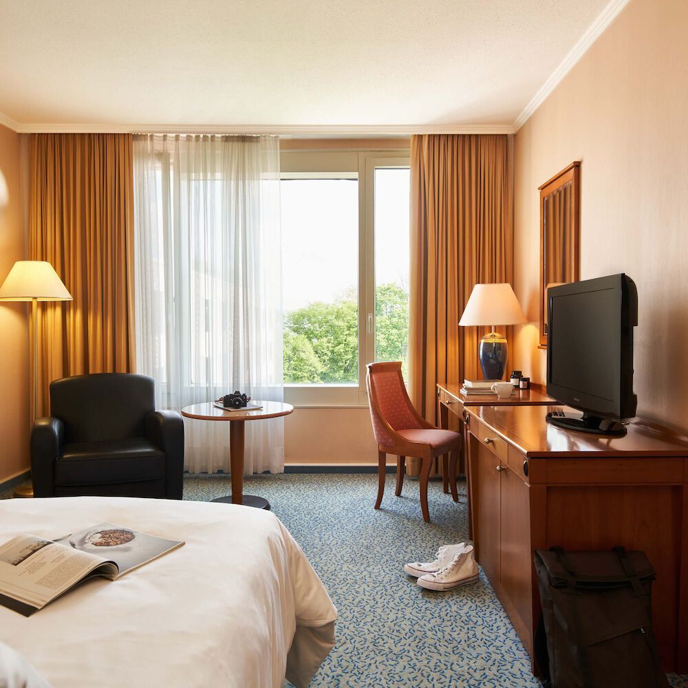 Hotelzimmer mit Bett, Schreibtisch, Stühlen und Fenster mit Vorhängen; hellblauer Teppich.