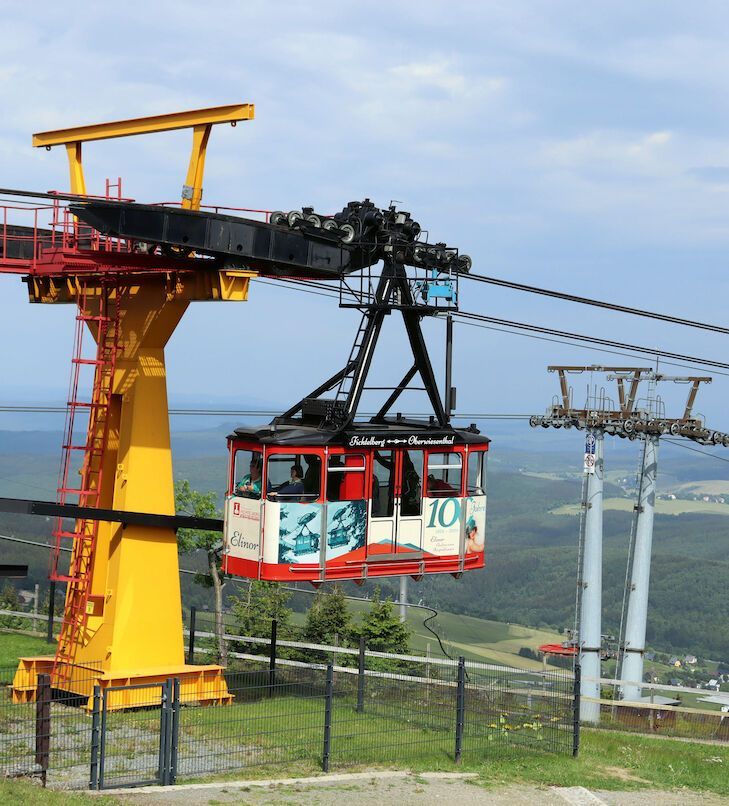 Seilbahn mit rot-weißer Kabine, die einen Berg mit gelber Stützkonstruktion hinaufführt.