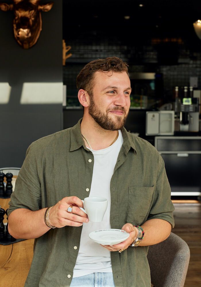 Ein Mann hält eine Kaffeetasse in der Hand, blickt zur Seite, trägt ein grünes Hemd und sitzt in einem Innenraum.