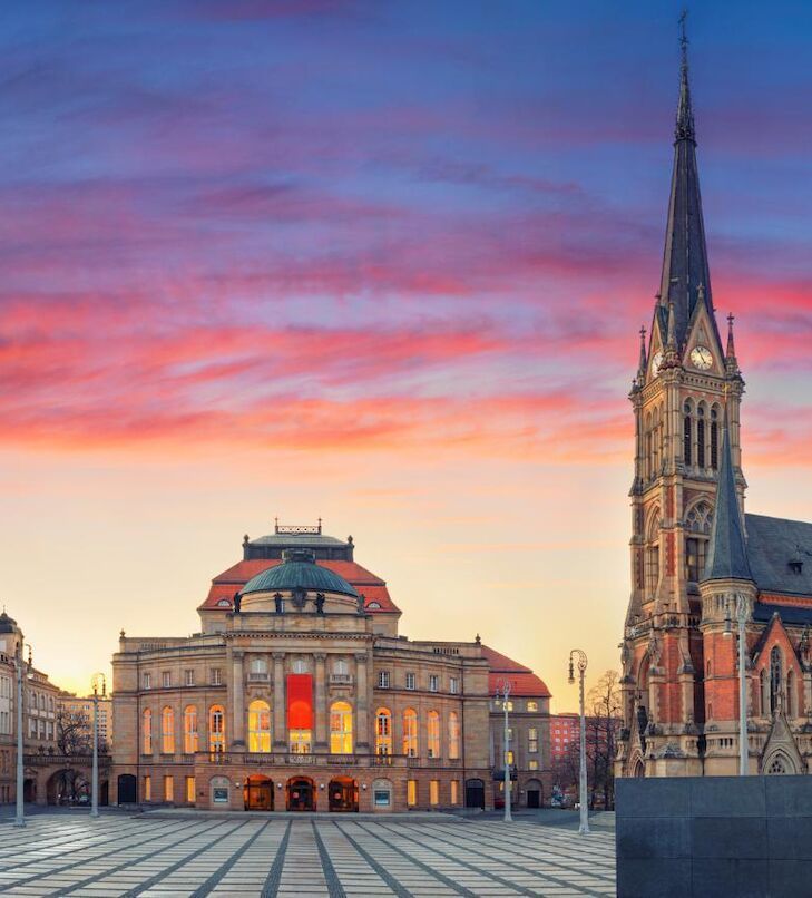 Theater- und Kirchengebäude auf einem Stadtplatz bei Sonnenuntergang; rosafarbener und orangefarbener Himmel.