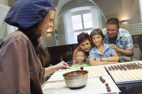 Eine Frau stellt Pralinen her, die einer Familie präsentiert werden. Die Familie beobachtet sie lächelnd in einem Laden.