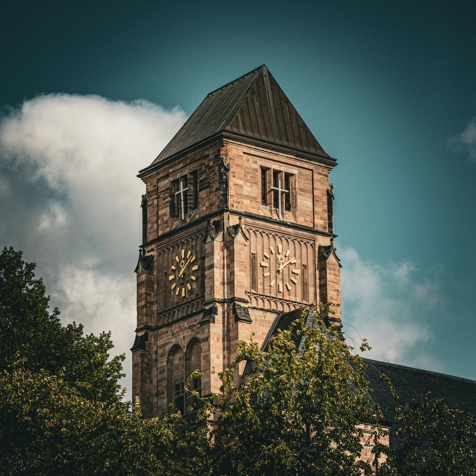 Steinturm mit Zifferblättern, gekrönt von einem dunklen Dach, eingerahmt von grünen Bäumen vor blauem Himmel.