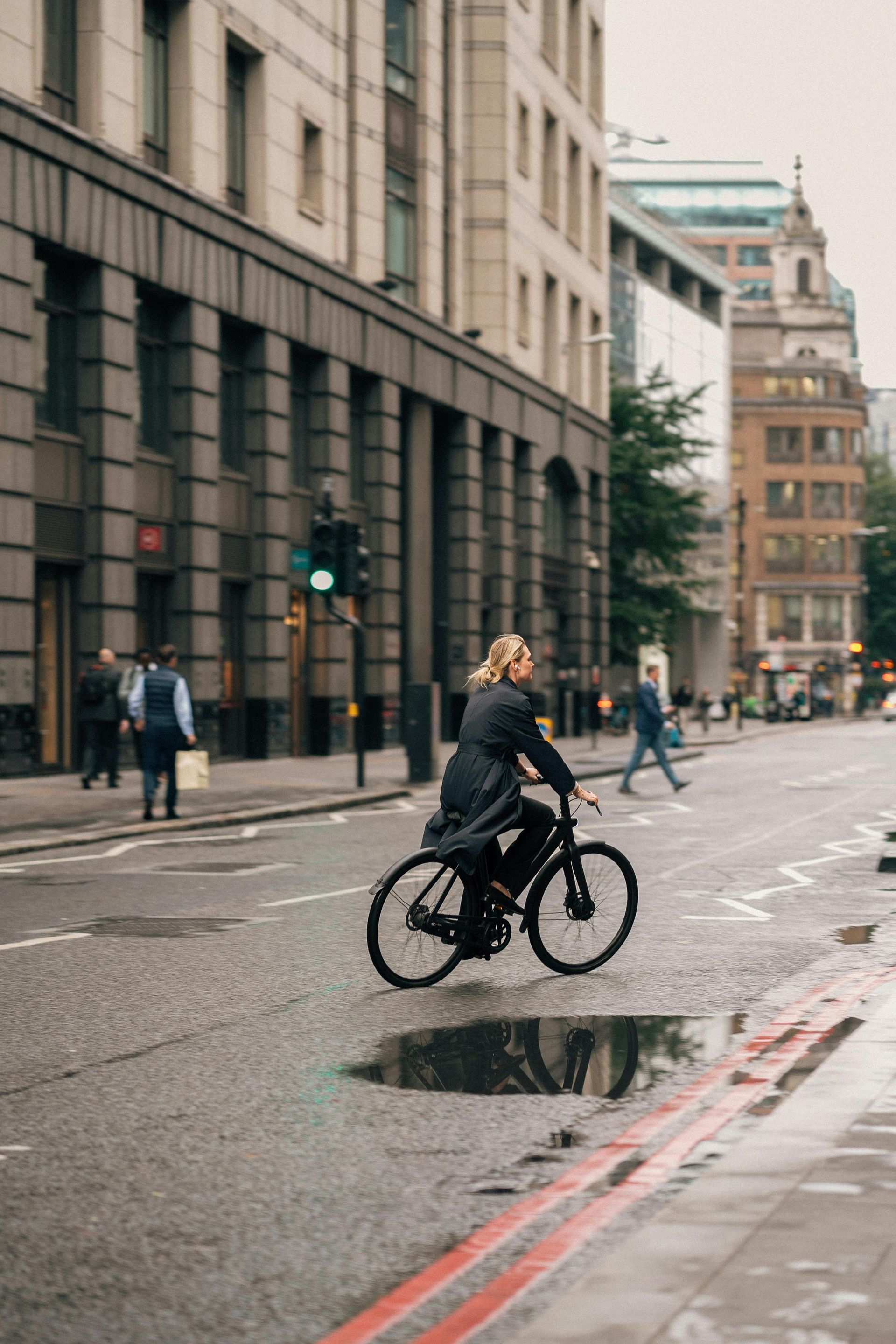 Eine Person fährt mit dem Fahrrad auf einer nassen Stadtstraße; im Hintergrund sind Gebäude zu sehen.