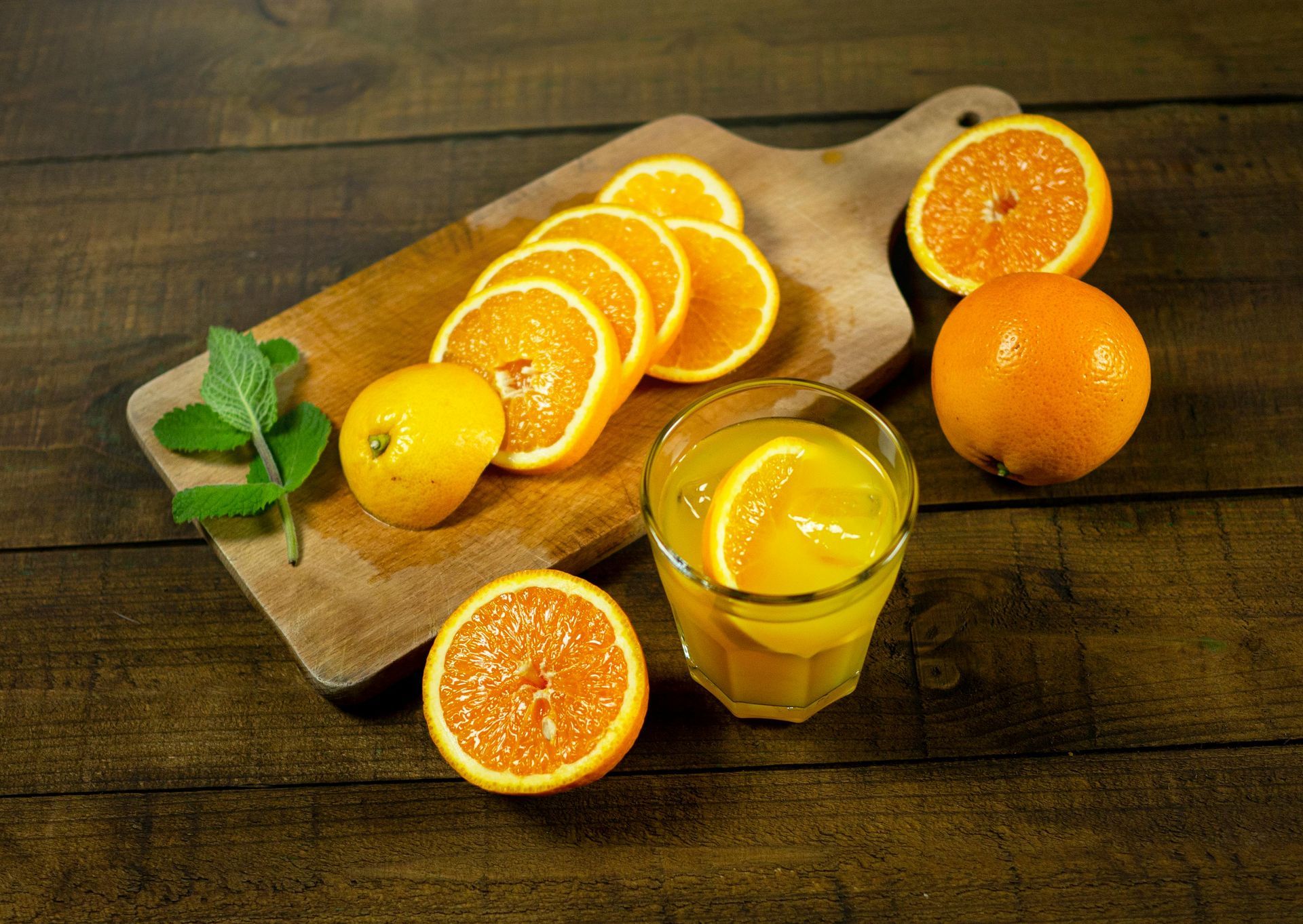 Ein Glas Orangensaft neben Orangen und einem Holzbrett, auf dem weitere Orangenscheiben liegen.