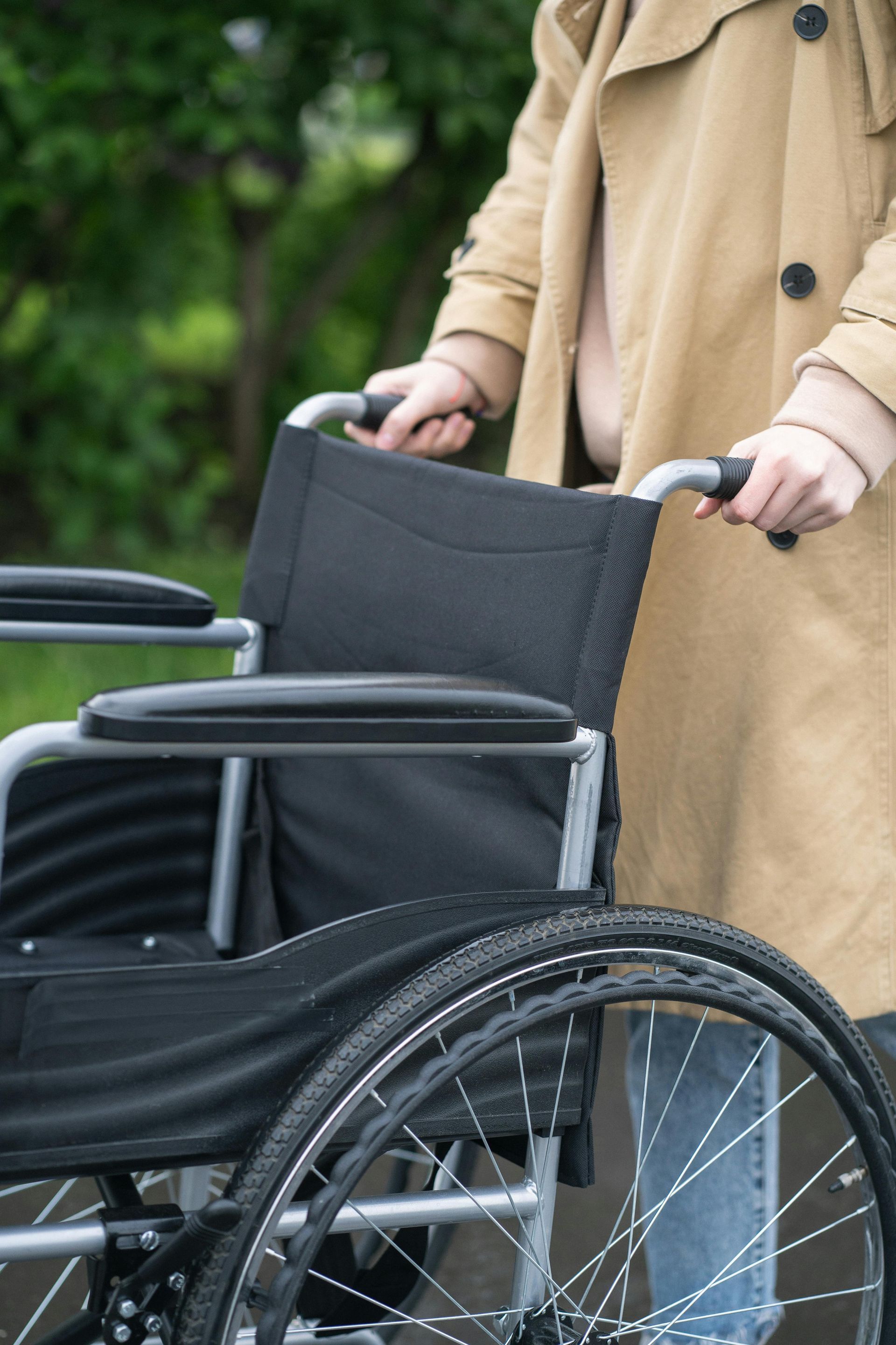 Person schiebt einen Rollstuhl im Freien; sie trägt einen beigen Mantel und einen schwarzen Rollstuhlsitzbezug.