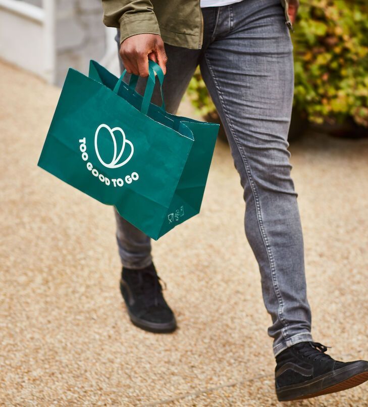 Eine Person geht, die eine grüne Tasche mit der Aufschrift „Too Good To Go“ trägt und graue Jeans und schwarze Schuhe anhat.