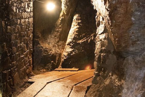 Schmaler, schwach beleuchteter Steintunnel mit Holzsteg und hellem Deckenlicht.