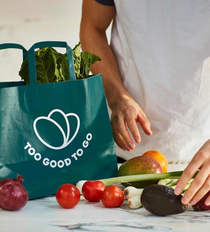 Eine Person packt Lebensmittel aus, darunter eine Too Good To Go-Tasche mit Logo; Gemüse liegt auf einer Theke.