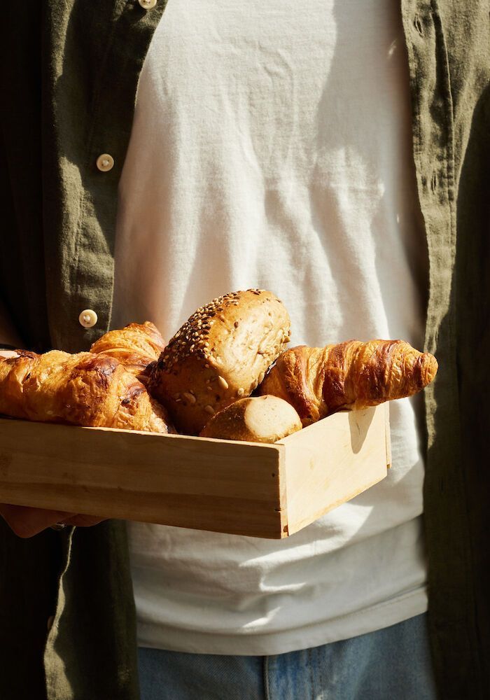 Eine Person hält ein Holztablett mit verschiedenen Backwaren, darunter Croissants und ein Brotlaib.