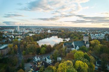 Luftaufnahme einer Stadt mit See, Bäumen, Gebäuden und einer Kirche. Bewölkter Himmel in der Abenddämmerung.