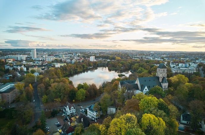 Luftaufnahme aus Chemnitz: Herbstbäume, ein See, eine Steinkirche sind zu sehen bei einem bewölkten Sonnenuntergangshimmel.