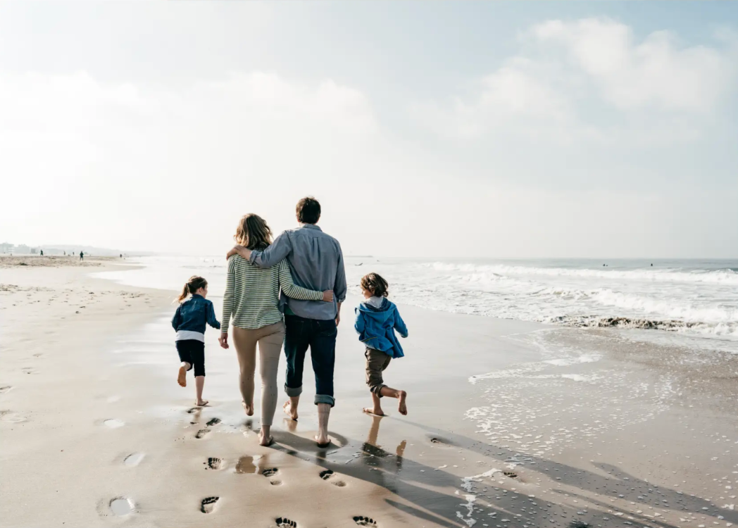 Sandstrand mit hohem Gras im Vordergrund, Meer und untergehende Sonne im Hintergrund.