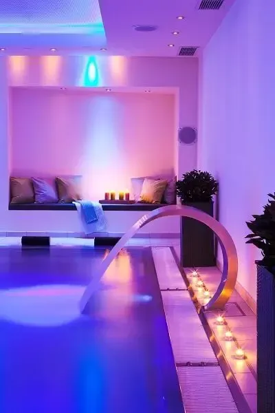 Indoor-Spa mit blauem Pool, Springbrunnen, Kerzen und einer in violettes und blaues Licht getauchten Bank.