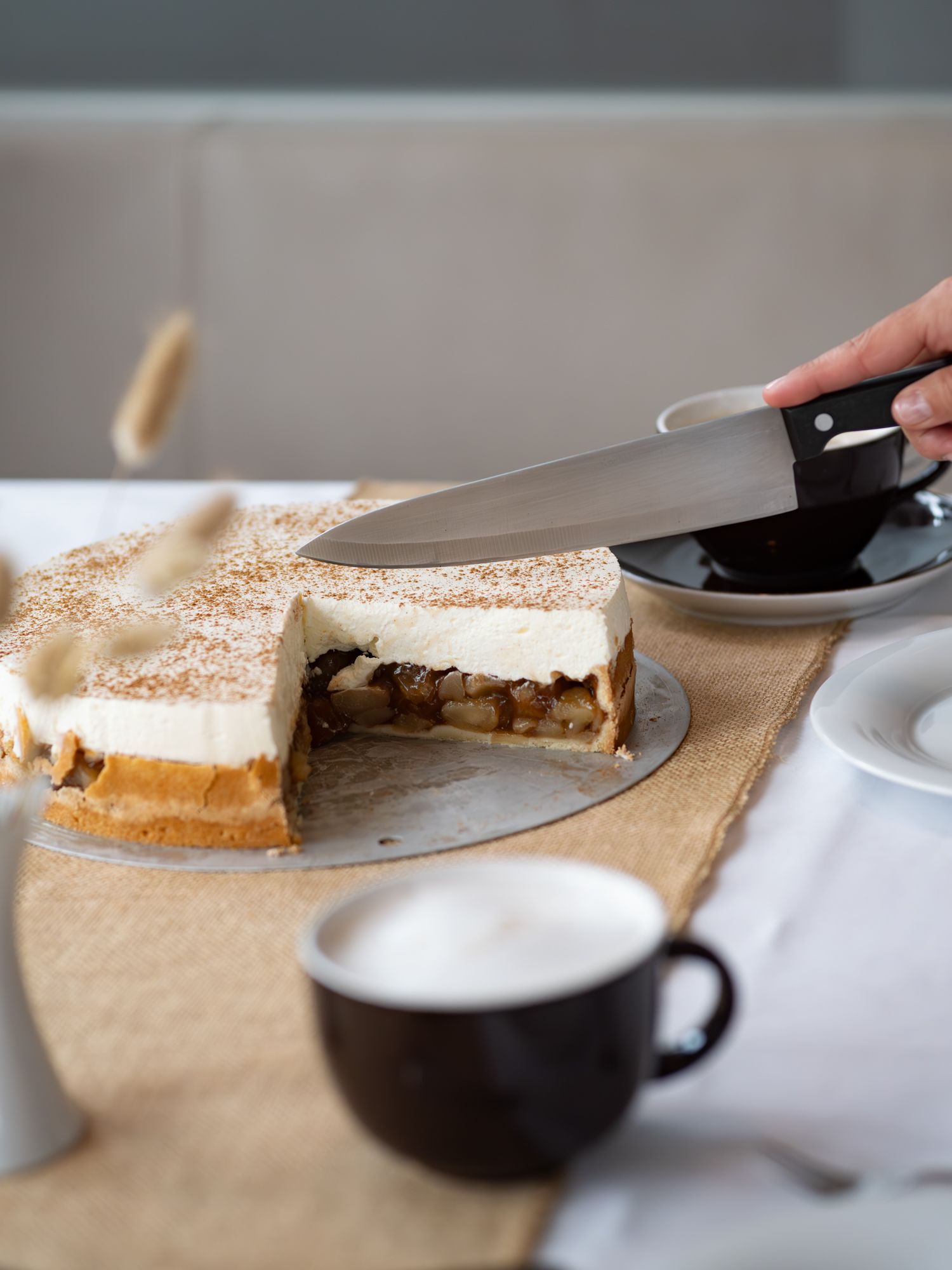 Eine Hand hält ein Messer über einen teilweise aufgeschnittenen Apfelkuchen mit Schlagsahne, der auf einem Tisch mit Kaffeetassen steht.