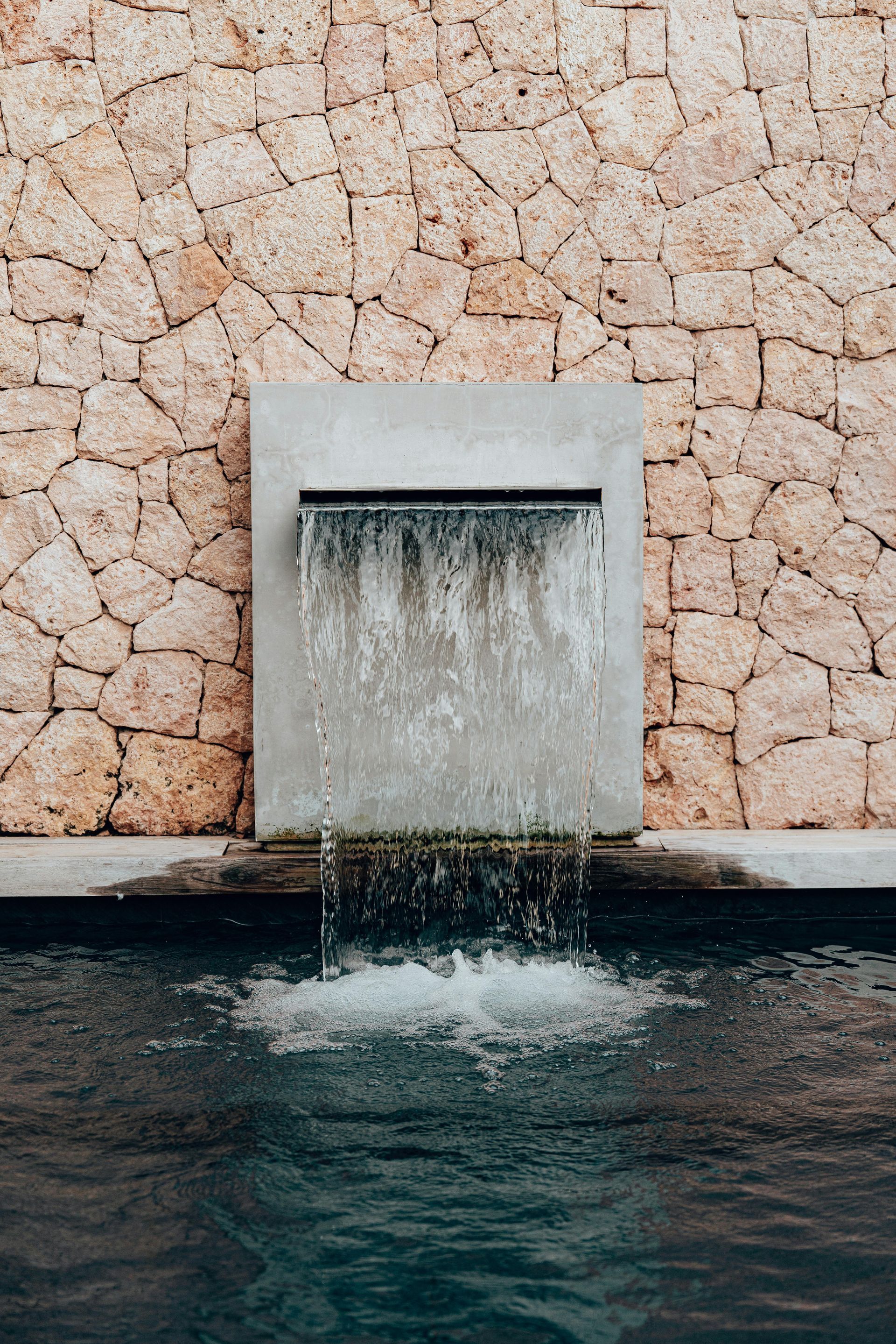 Wasserfall, bei dem das Wasser von einem Betonplatz in einen dunklen Pool vor einer Steinmauer stürzt.