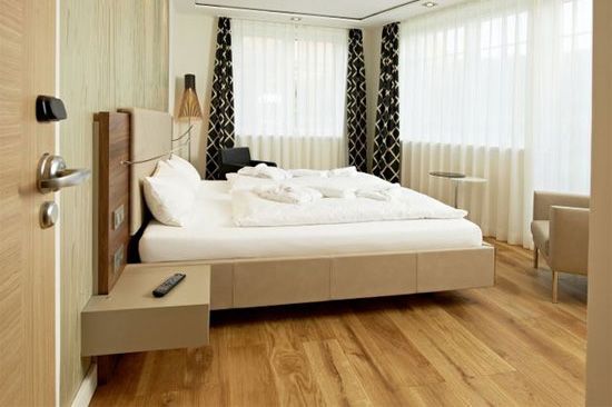 Schlafzimmer mit Bett, Nachttisch und Stuhl. Neutrale Farben mit hellem Holzboden und weißen Vorhängen.