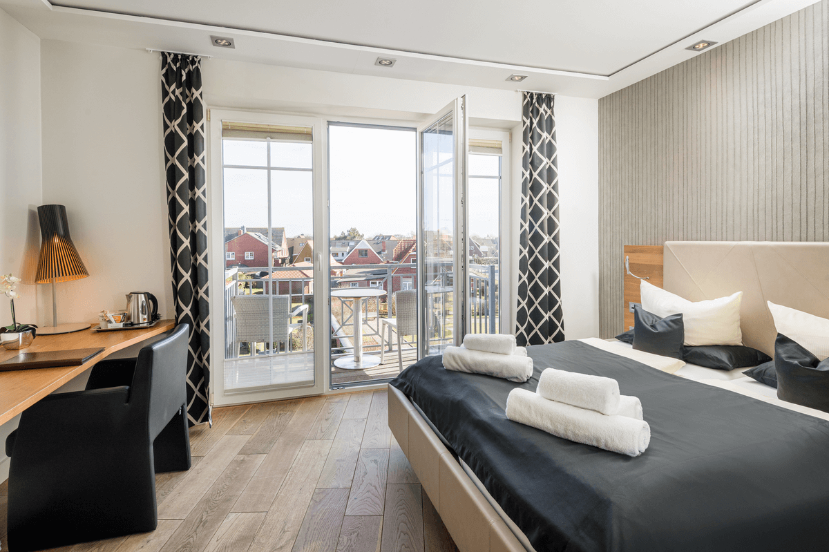 Schlafzimmer mit Bett, Schreibtisch, Balkon und offenen Doppeltüren.