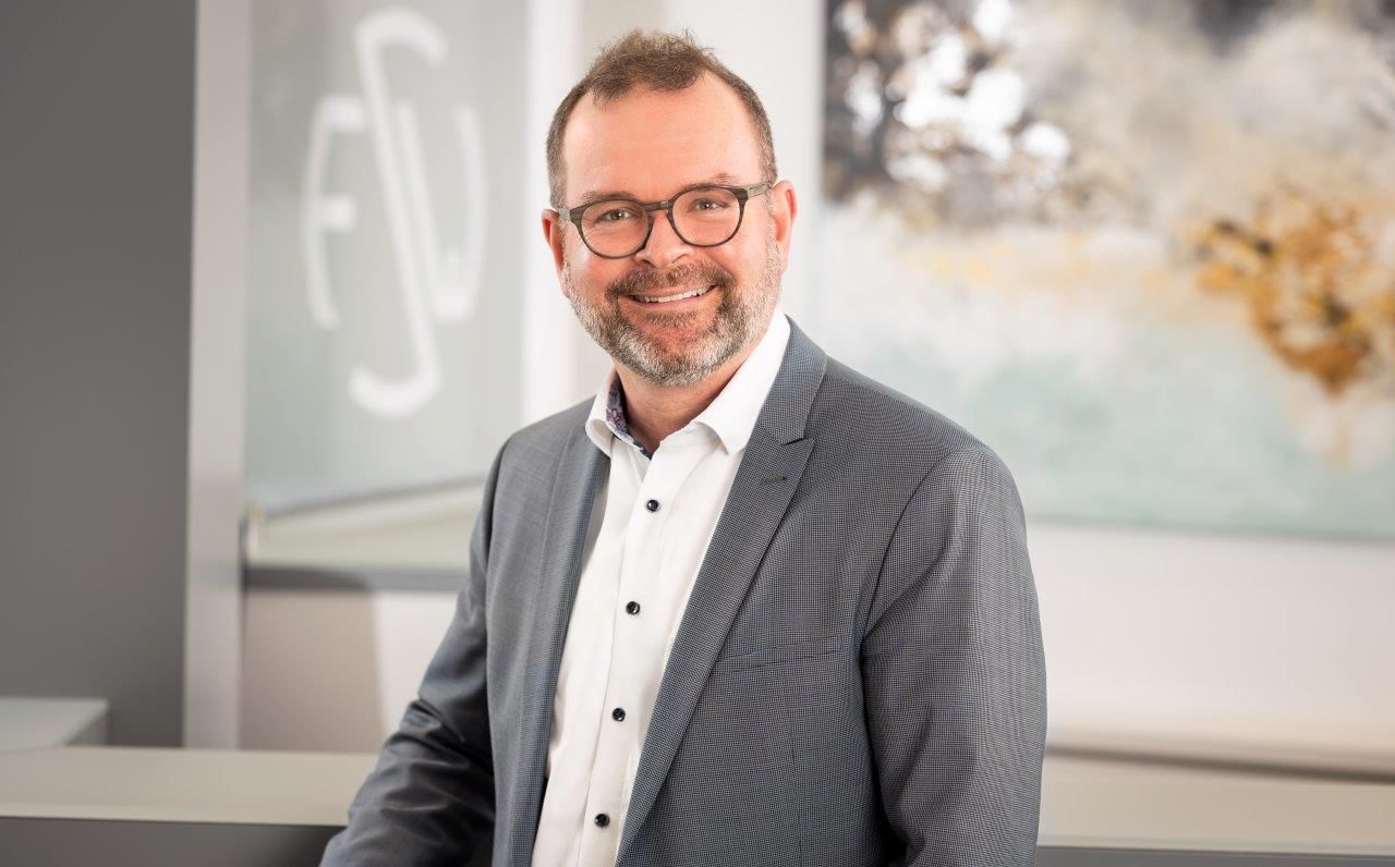 Ein Mann in Anzug und Brille lächelt vor einem Gemälde.