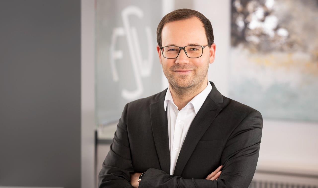 Ein Mann in Anzug und Brille steht mit verschränkten Armen da.