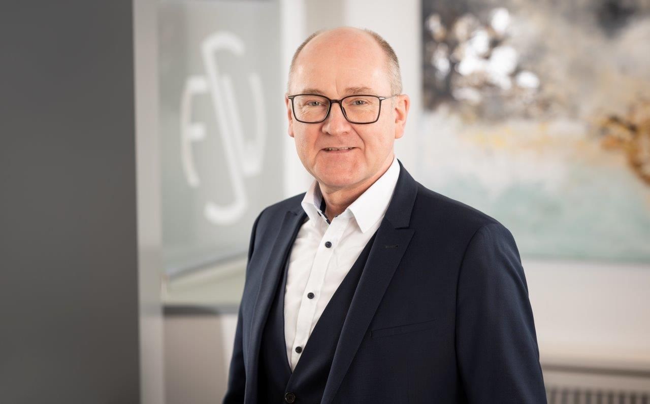 Ein Mann in Anzug und Brille steht vor einem Gemälde.