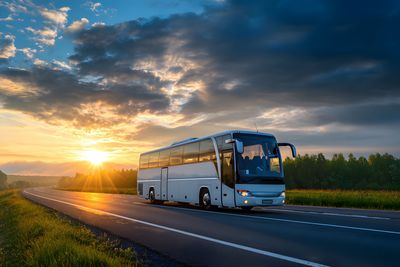 Weißer Reisebus fährt bei Sonnenuntergang auf einer Straße mit goldenem Licht und bewölktem Himmel.