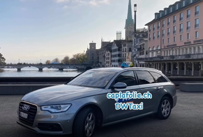Silbernes Audi-Taxi mit „DW Taxi“-Logo in Zürich, Schweiz, vor einer Brücke und Gebäuden.