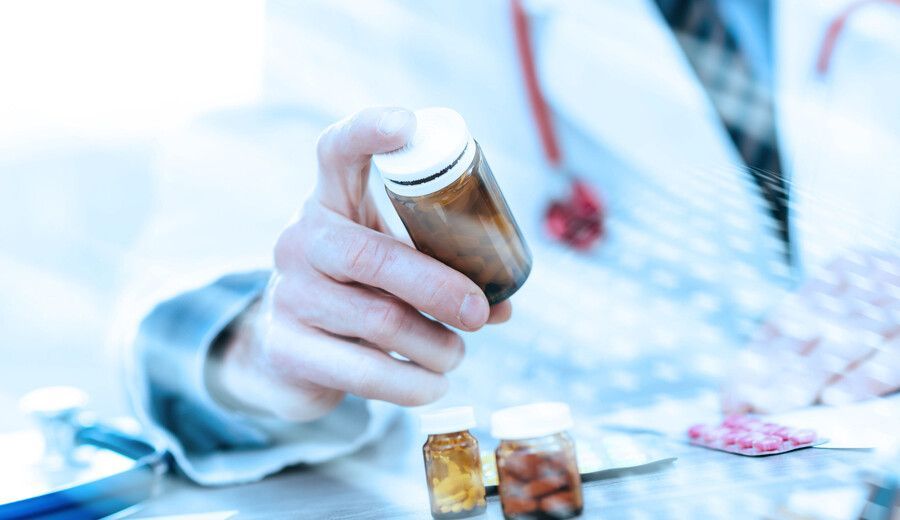 Una mano sostiene un frasco de pastillas recetadas sobre un formulario médico borroso con cápsulas dispersas.