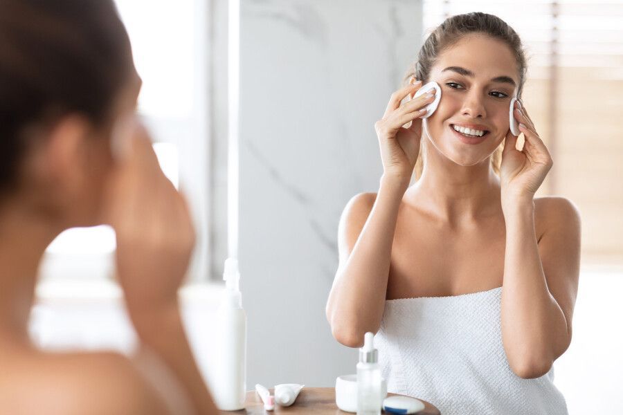 Mujer aplicándose crema facial frente al espejo del baño, sonriendo mientras usa discos de algodón.
