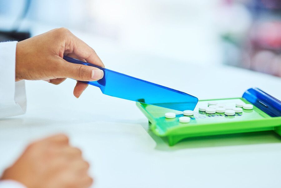 Una mano coloca una cuchara azul en un pastillero verde con tabletas blancas sobre una mesa.
