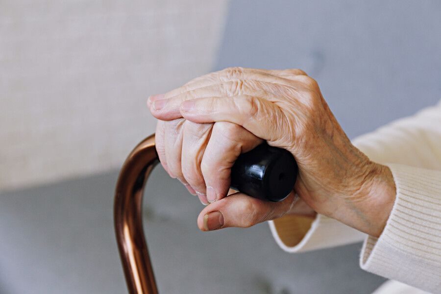 Manos sujetando el mango negro de un bastón de madera curvado sobre un sofá gris.