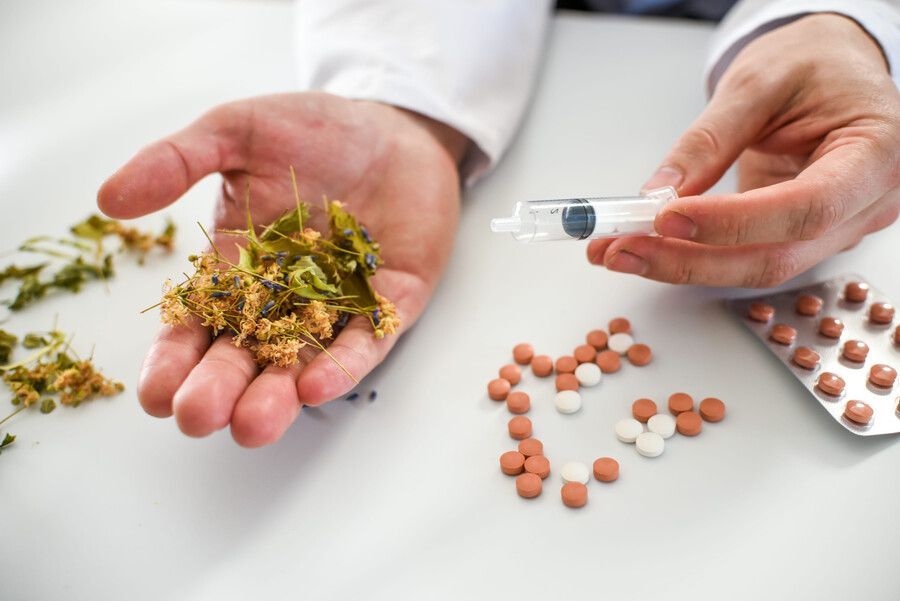 Manos sosteniendo un suplemento herbal y una jeringa junto a pastillas esparcidas sobre una mesa blanca.