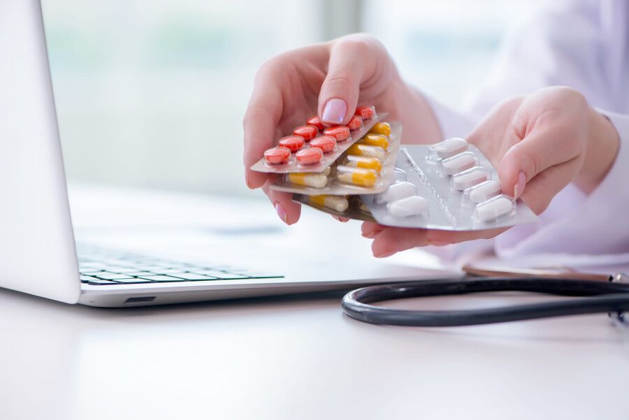 Manos sosteniendo blísteres de pastillas cerca de una computadora portátil y un estetoscopio sobre un escritorio.