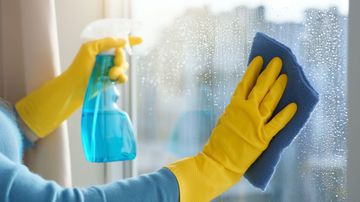 Une personne nettoie une fenêtre avec des gants jaunes, un chiffon bleu et un flacon pulvérisateur bleu.