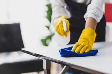 Une personne portant des gants jaunes essuie une table avec un chiffon bleu dans un bureau.