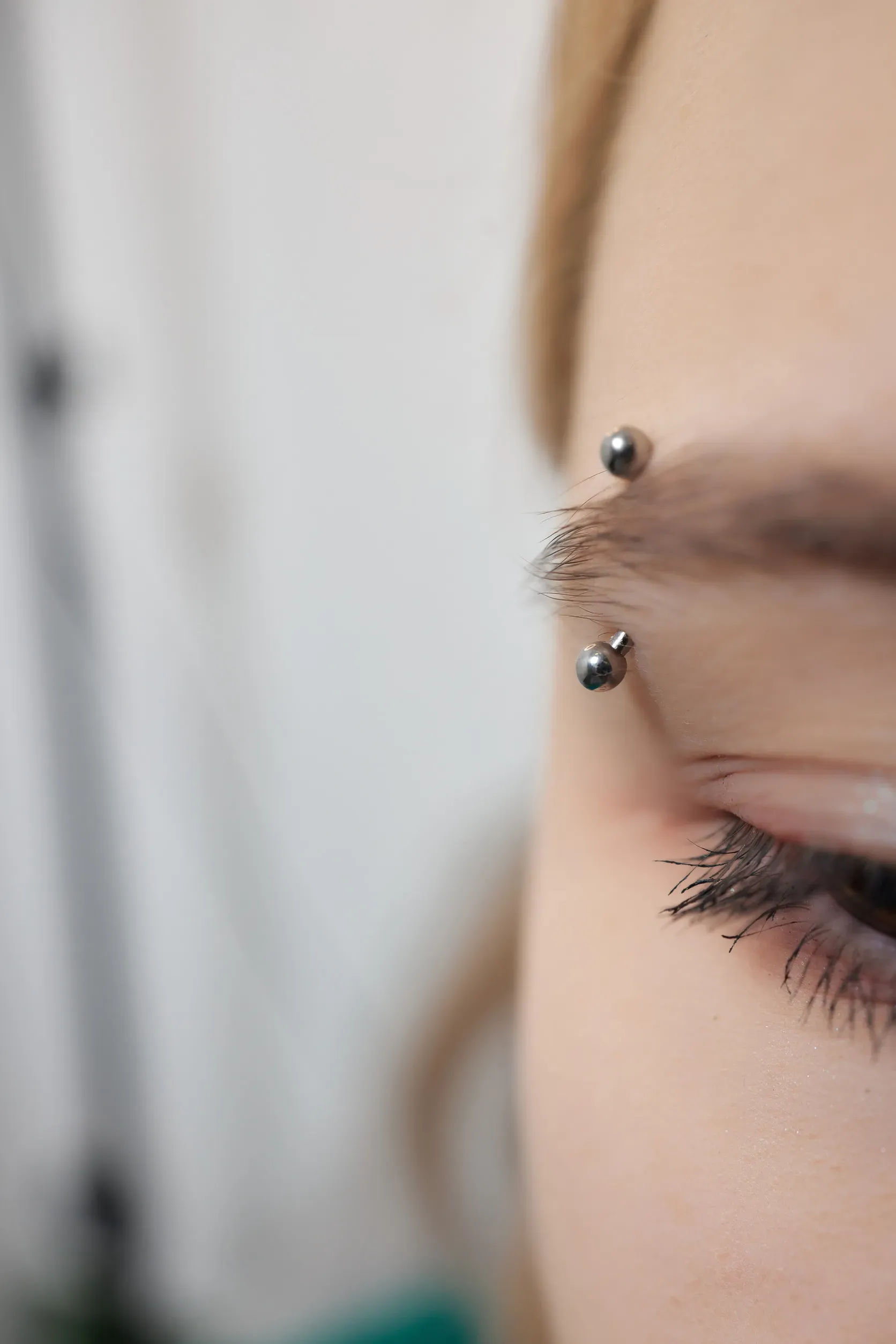 Primer plano del ojo y la ceja de una persona con dos piercings. La piel está pálida y el fondo está borroso.