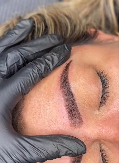 Mano enguantada sosteniendo la piel cerca de la ceja de una persona con un nuevo tatuaje cosmético.