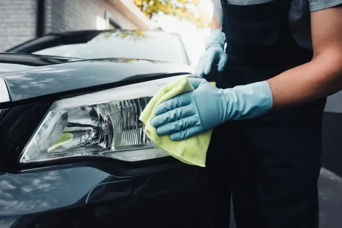 Persona que lleva guantes azules limpiando un faro de un coche con un paño amarillo.
