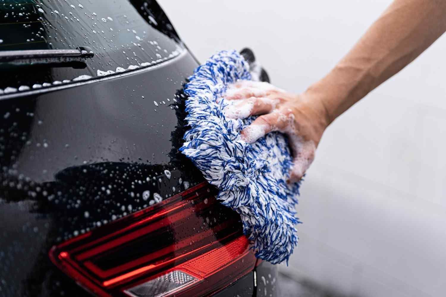 Persona lavando un coche negro con un guante de lavado azul y blanco, con espuma visible.