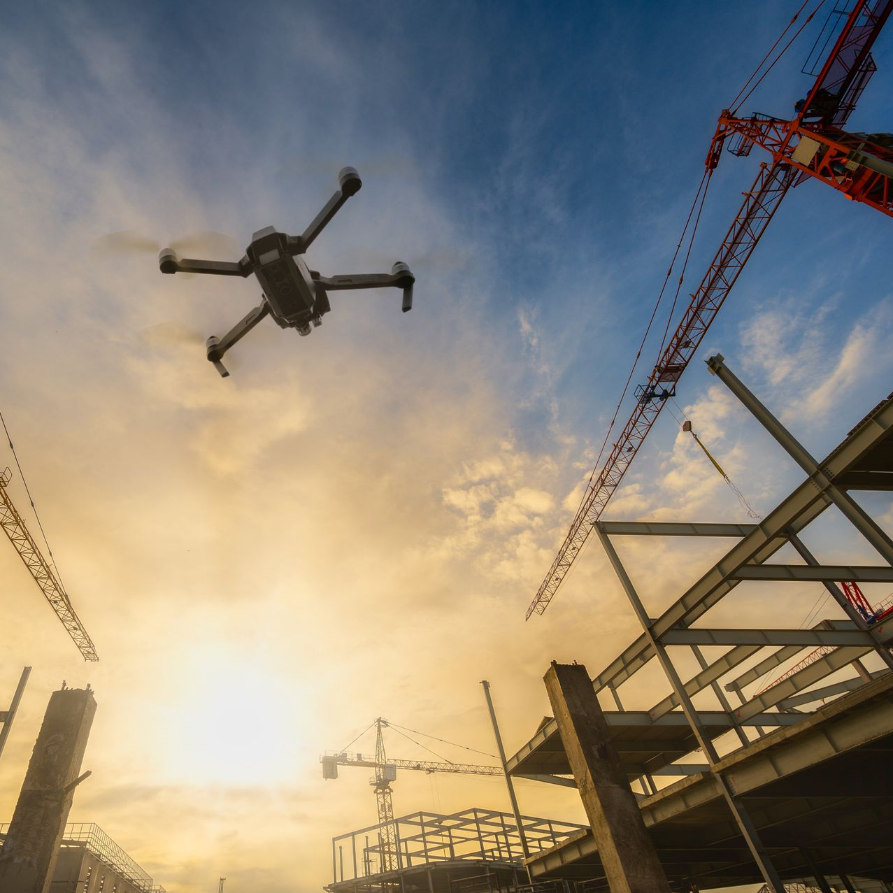 Survol aérien d'un chantier au coucher du soleil, avec des grues et une structure métallique