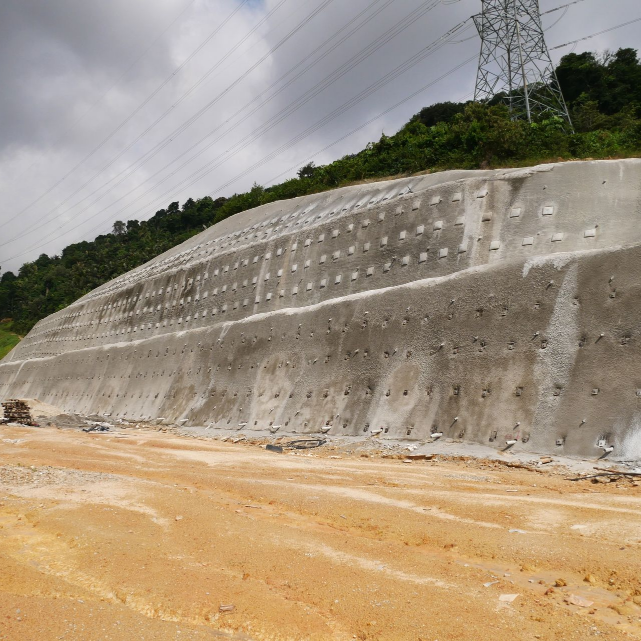 Mur de soutènement en béton au bord d'un chemin de terre, avec des collines et des lignes électriques sous un ciel nuageux