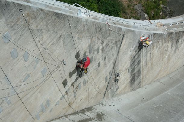 Des ouvriers casqués escaladent la pente abrupte d'un barrage en béton à l'aide de cordes et d'équipements de sécurité
