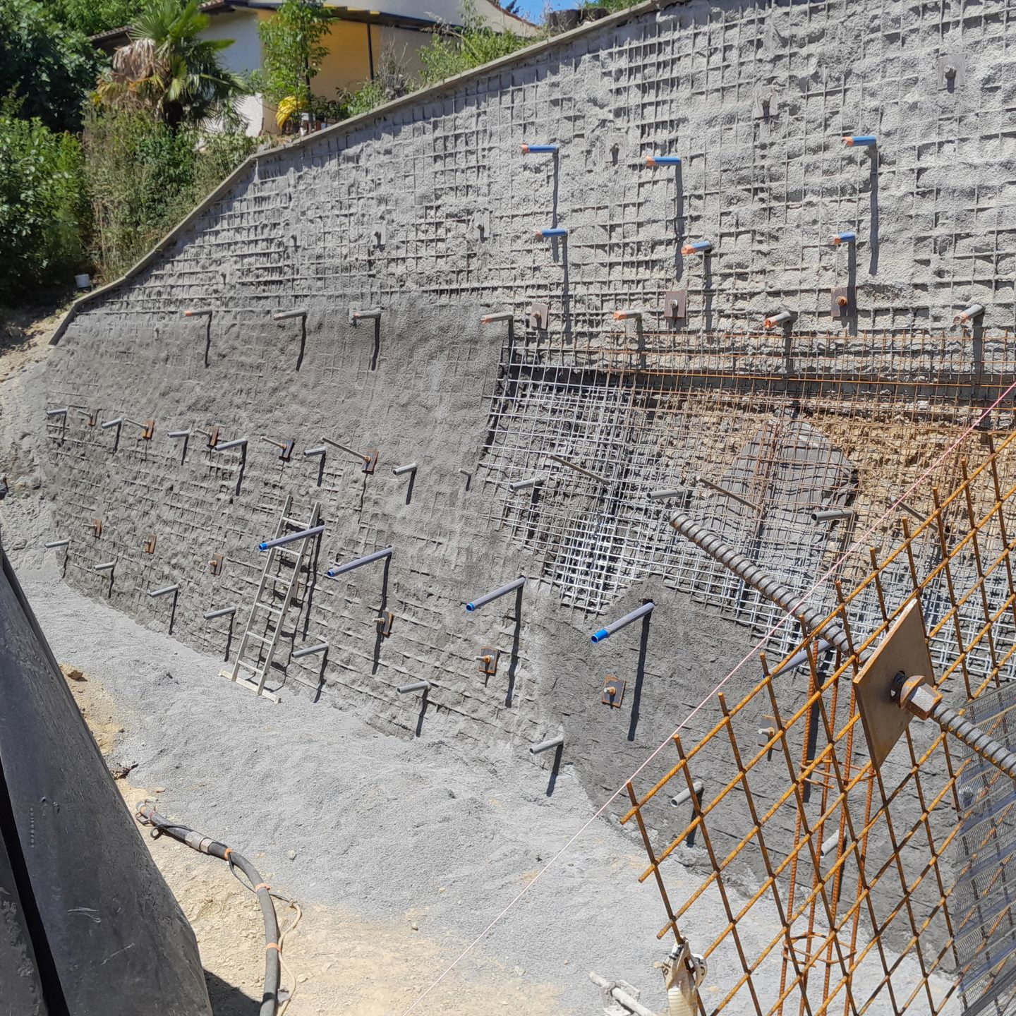 Mur de soutènement en béton en terrasses avec ancrages bleus et treillis métallique sur un versant en pente