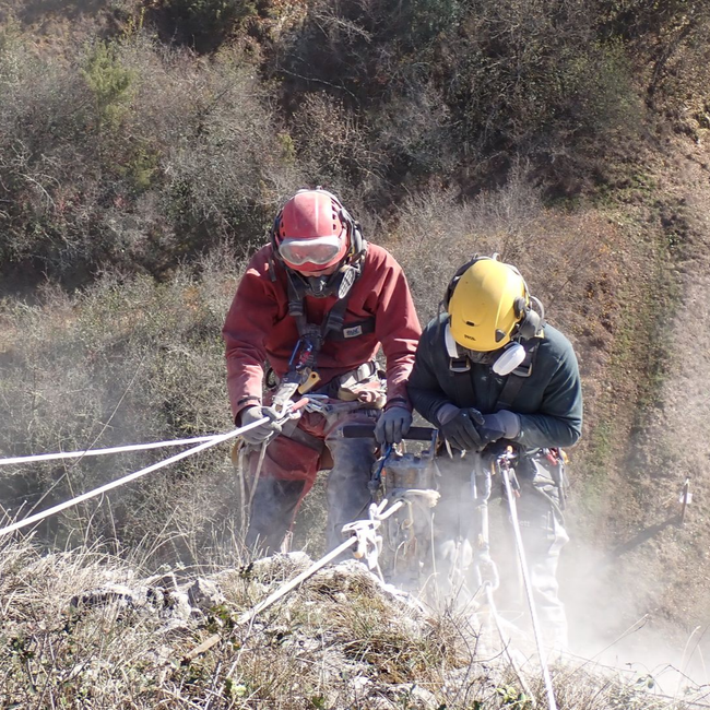 Deux alpinistes casqués et harnachés descendent en rappel une pente rocheuse, entourés de cordes et de poussière
