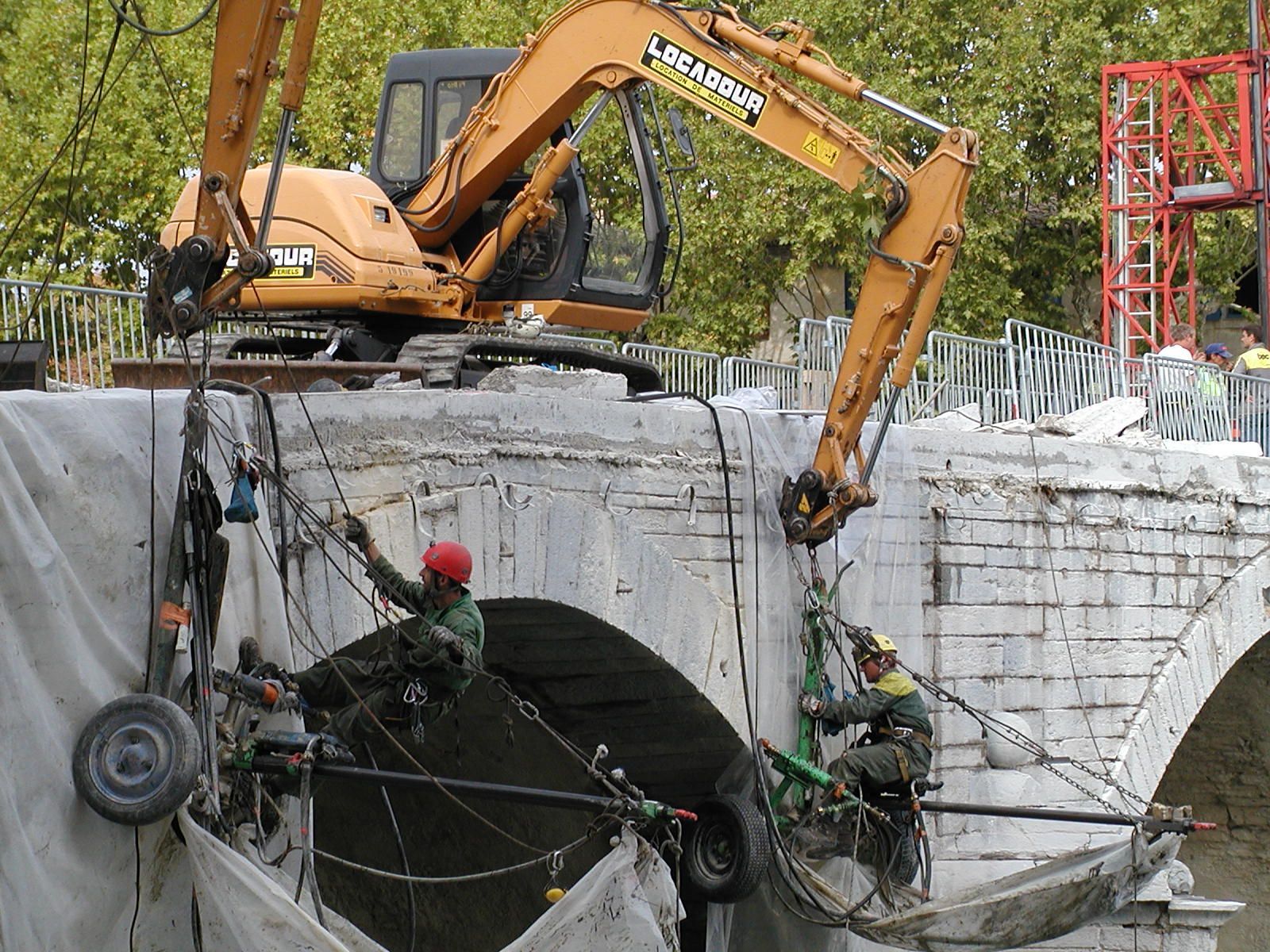 Une excavatrice démolit un pont en béton tandis que des ouvriers sécurisent les câbles en contrebas sur un chantier de construction